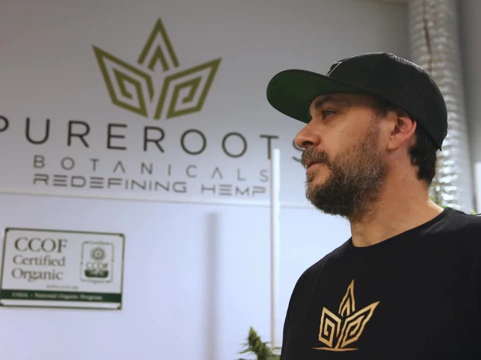 Ben Cerullo in the Pure Roots grow room, in front of the Pure Roots Botanicals sign and the CCOF Certified Organic sign