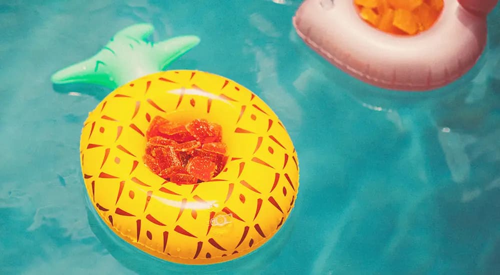 Pure Roots Botanicals Delta-9 Gummies infused with Live Rosin floating in a pineapple-shaped pool float, with a flamingo float in the background, surrounded by clear blue water, a fun summer vibe.