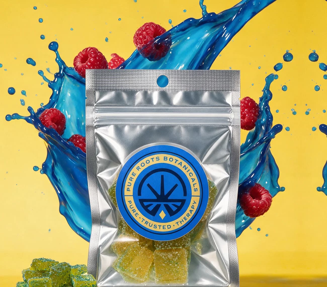 A silver Pure Roots Blue Raspberry and Green Apple gummies pouch in front of a blue fruit splash with raspberries, on a yellow backdrop