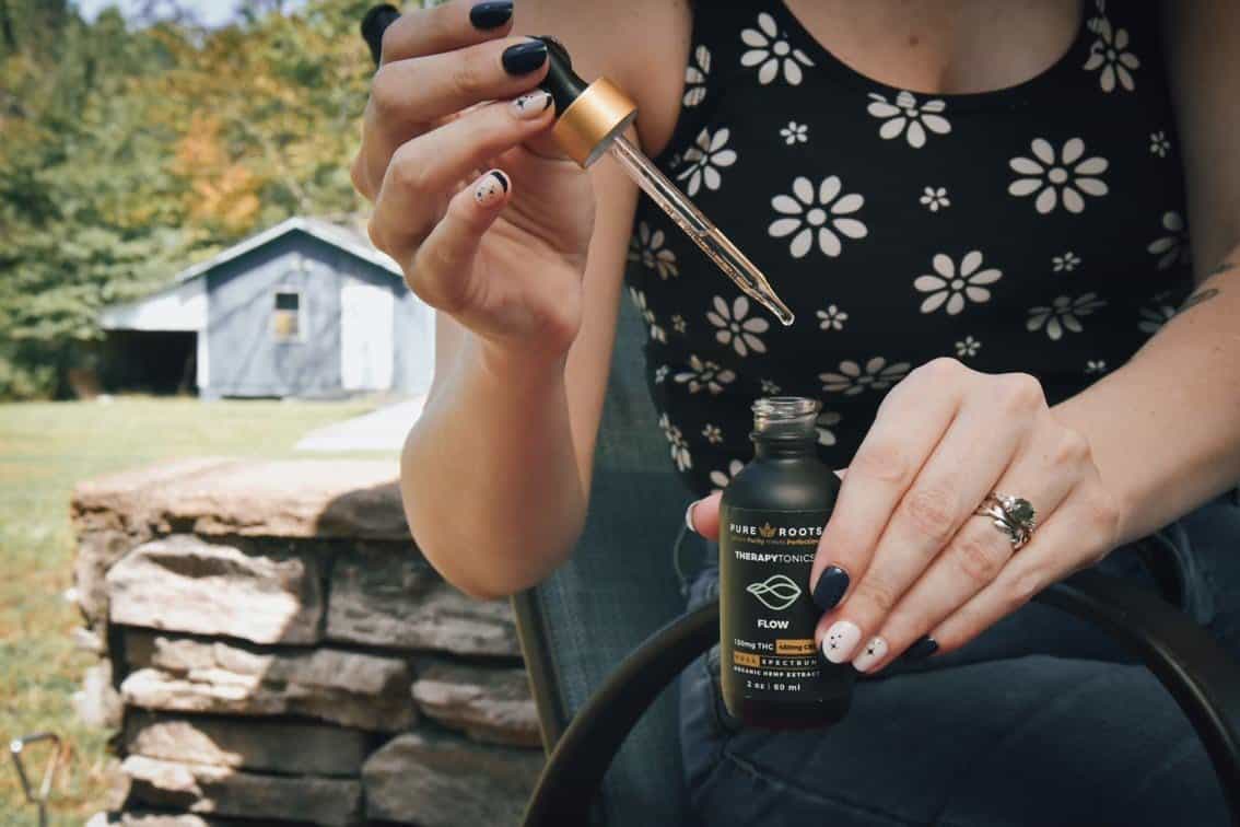 Lady holding a Pure Roots Botanicals CBD tincture bottle, pulling the dropper out of the bottle with oil visible inside the dropper.